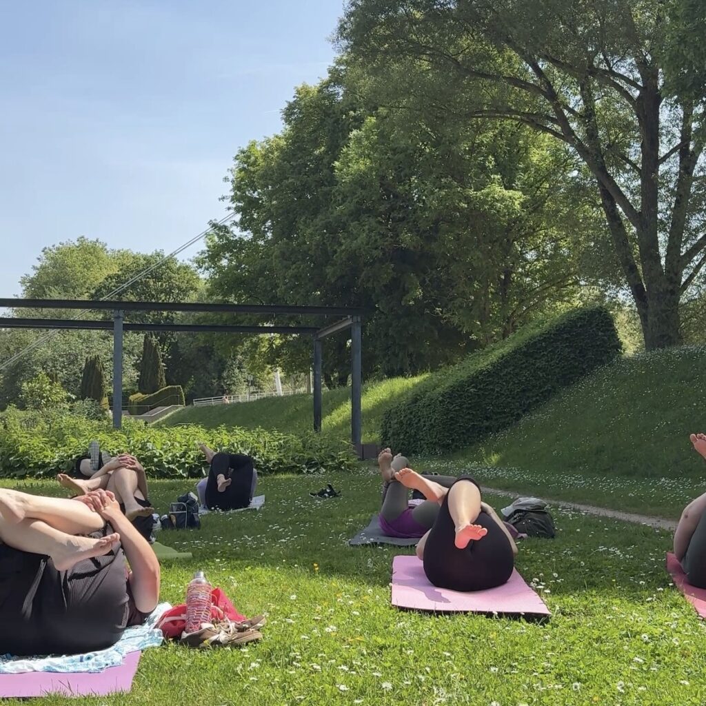 Teilnehmer praktizieren die Yoga-Asana Sucirandhrasana im Enzauenpark Pforzheim zur Dehnung der Hüfte und Entlastung des unteren Rückens